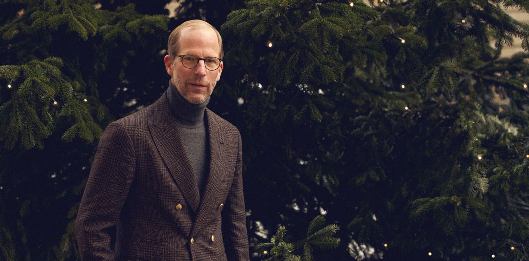 Mann mit Brille und braunem kariertem Mantel steht vor einem großen Weihnachtsbaum mit Lichterketten.