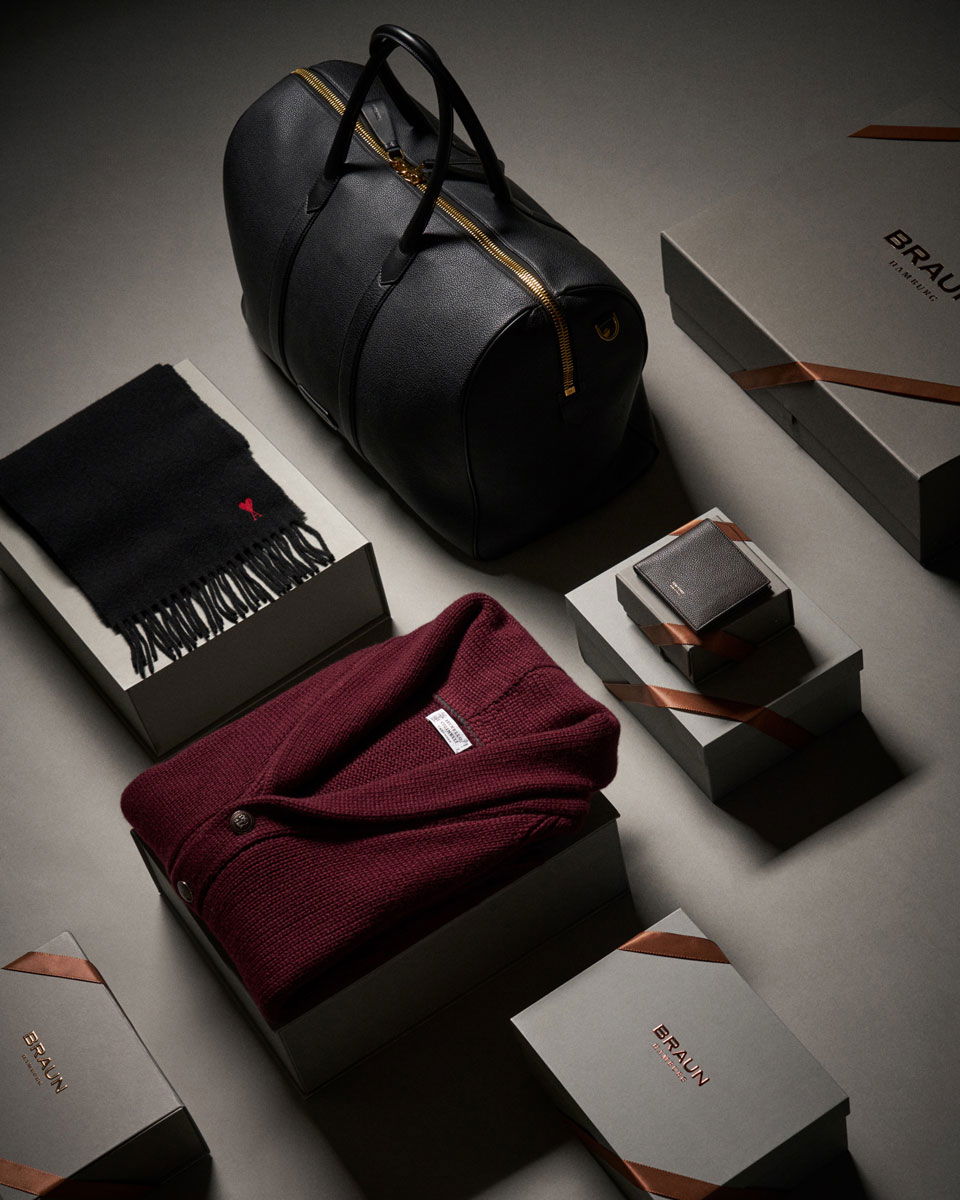 Schwarze Ledertasche, schwarzer Schal, weinroter Strickpullover, graue Geschenkboxen mit braunen Bändern und schwarzer Geldbörse.