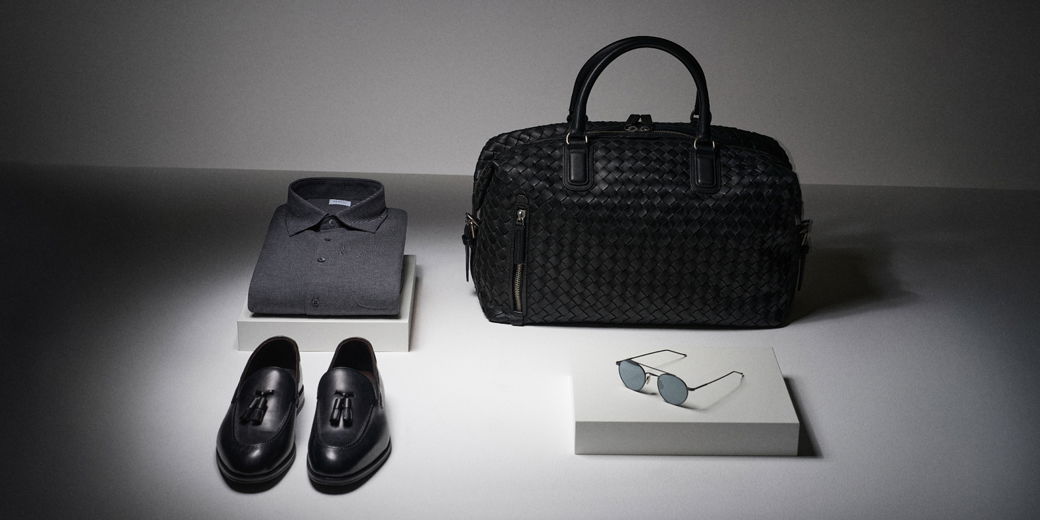 Folded dark gray polo shirt, black leather tassel loafers, woven black leather duffel bag, and round sunglasses on white boxes.