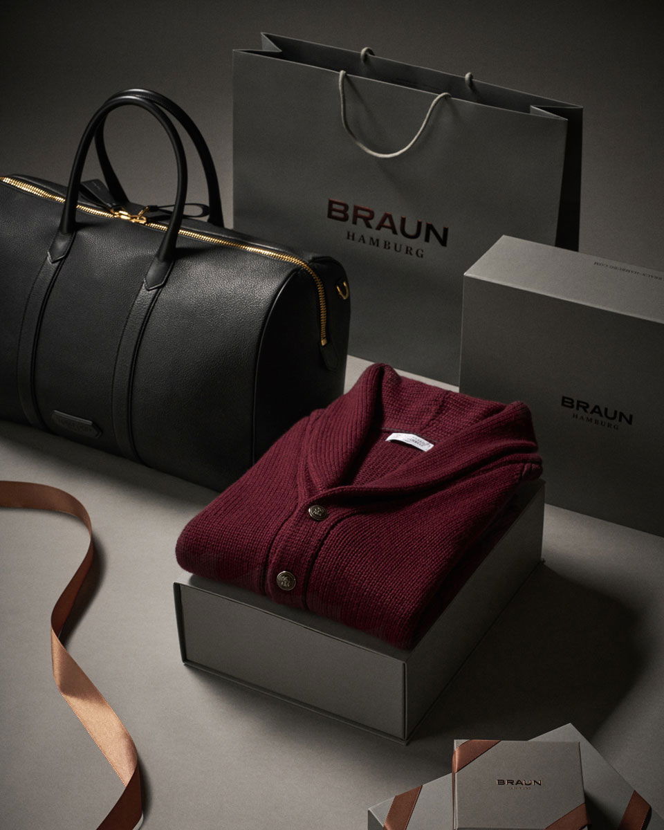 Schwarze Ledertasche, graue Geschenktüten und -boxen mit Braun Hamburg Logo, roter Strickpullover auf Box, braunes Geschenkband.
