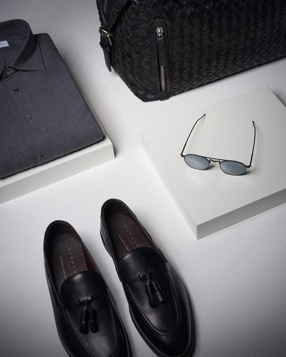 Folded dark gray dress shirt, woven black leather bag, black tassel loafers, and oval sunglasses on white platforms.