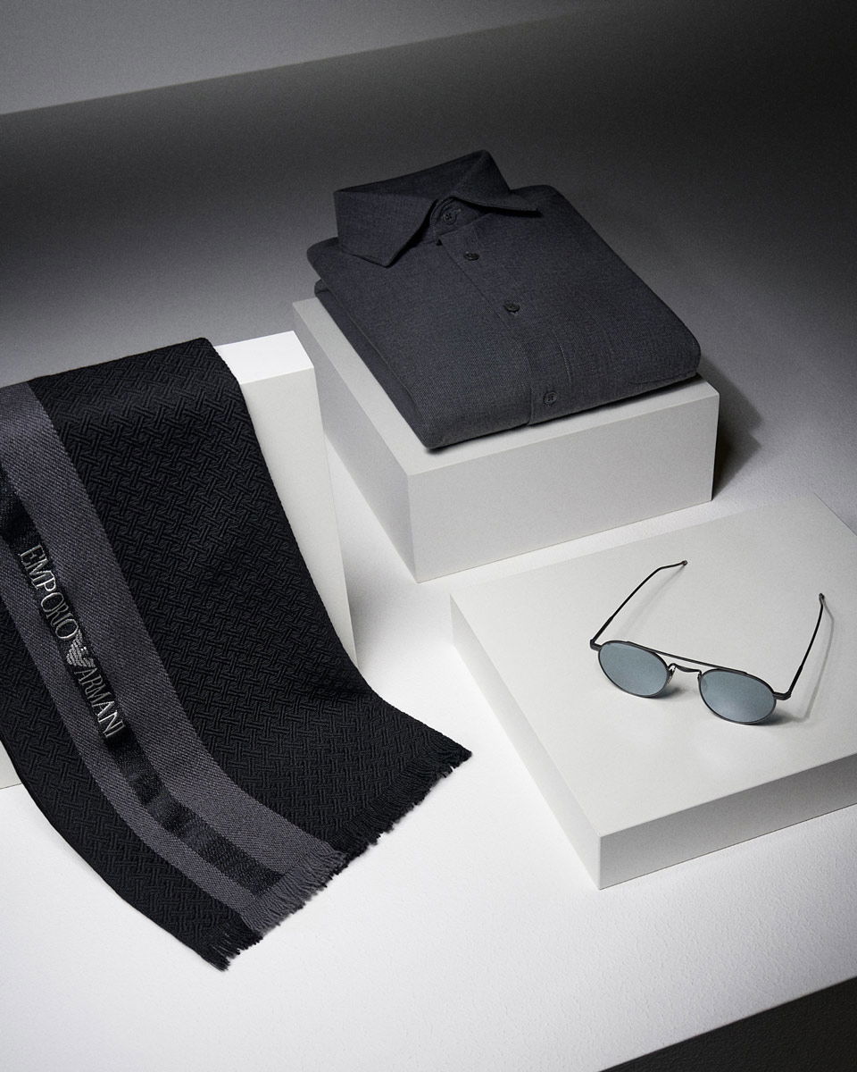 Schwarzer Emporio Armani Schal, gefaltetes dunkles Hemd und schwarze Sonnenbrille auf weißen Podesten arrangiert.