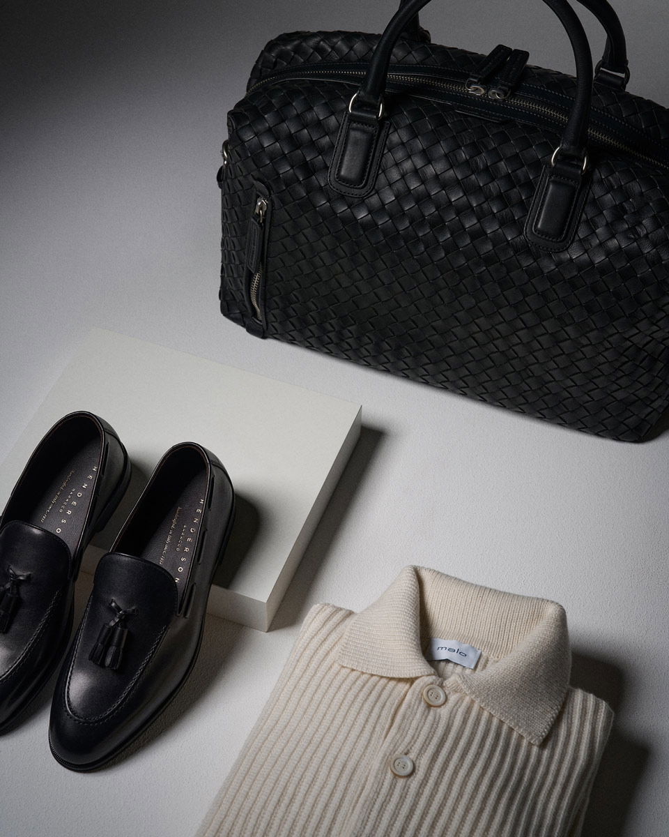 Schwarze geflochtene Lederreisetasche, schwarze Lederschuhe mit Quasten, cremefarbener Rippsweater mit Kragen auf grauem Untergrund.