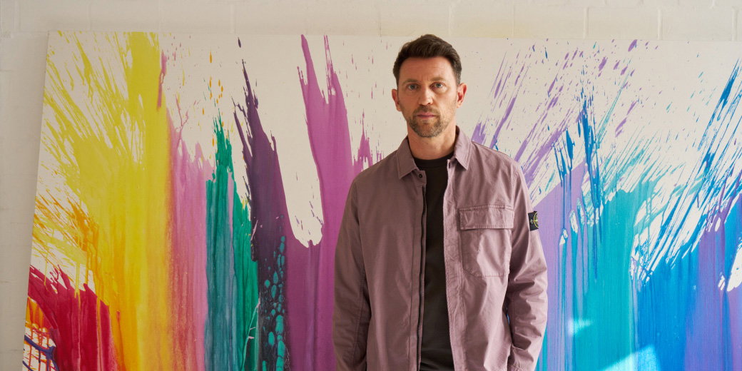 Man with short brown hair and beard wearing a mauve jacket and black shirt standing in front of colorful abstract paint splashes on white wall.
