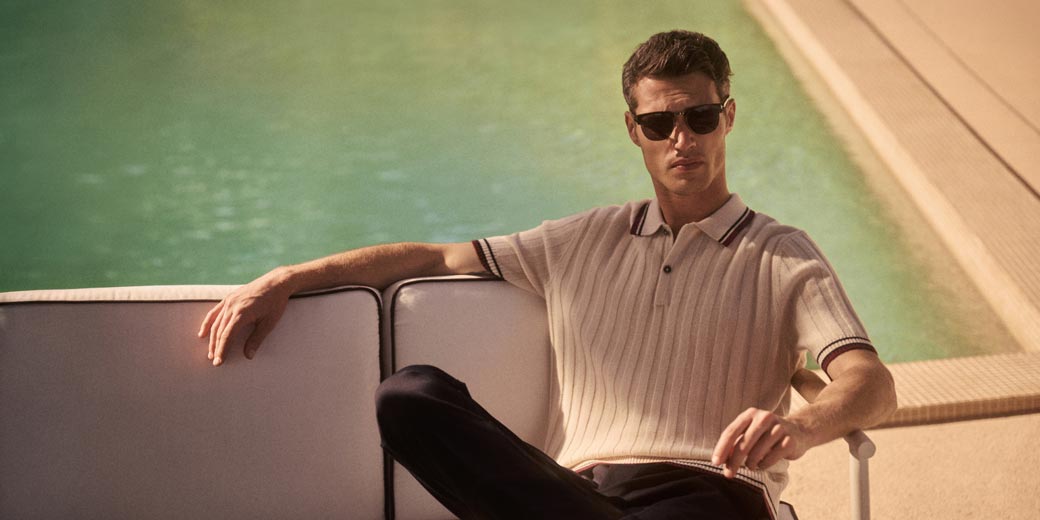 Man wearing sunglasses and a beige short-sleeve knit polo shirt sitting on a white outdoor sofa by a pool.