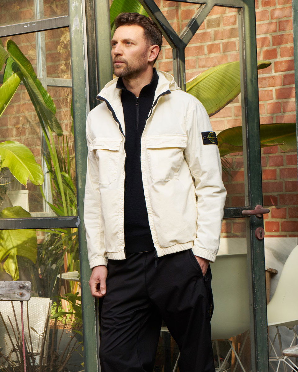Man wearing a white jacket and black pants stands indoors near a glass door with large green plants and brick wall background.