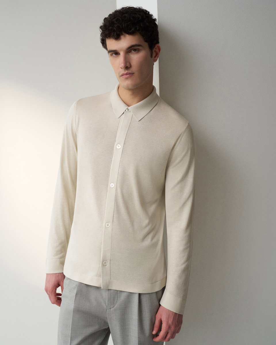 Young man with curly dark hair wearing a beige button-up long-sleeve shirt and light gray trousers, leaning against a wall. Young man with curly dark hair wearing a beige button-up long-sleeve shirt and light gray trousers, leaning against a wall.