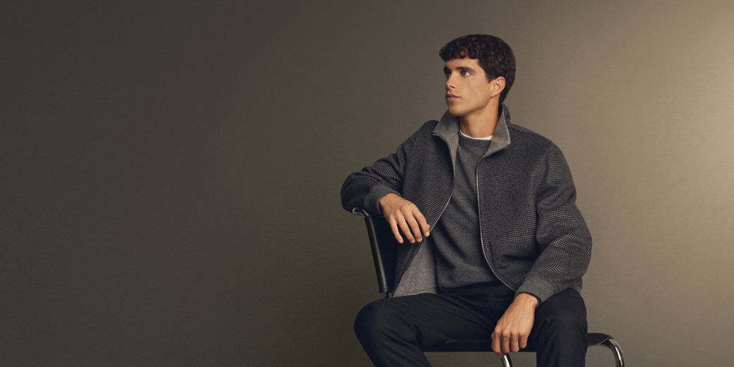 Junger Mann mit kurzen braunen Haaren sitzt seitlich auf Stuhl, trägt grauen Pullover und karierten grauen Mantel vor dunklem Hintergrund.