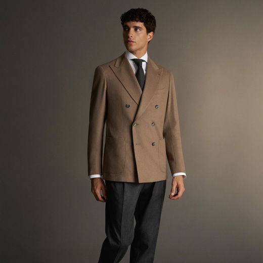 Junger Mann trägt braune doppelte Sakkojacke, weißes Hemd, schwarzen Schlips und graue Anzughose vor neutralem Hintergrund.