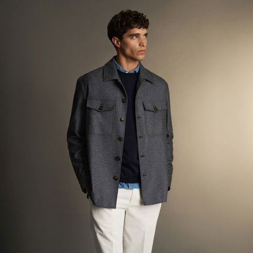 Junger Mann trägt graue Hemdjacke, blauen Pullover, hellblaues Hemd und weiße Hose vor schlichtem Hintergrund.