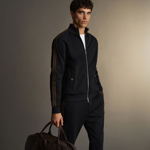 Junger Mann trägt schwarzen Trainingsanzug mit orangenen Streifen und hält eine braune Reisetasche vor neutralem Hintergrund.