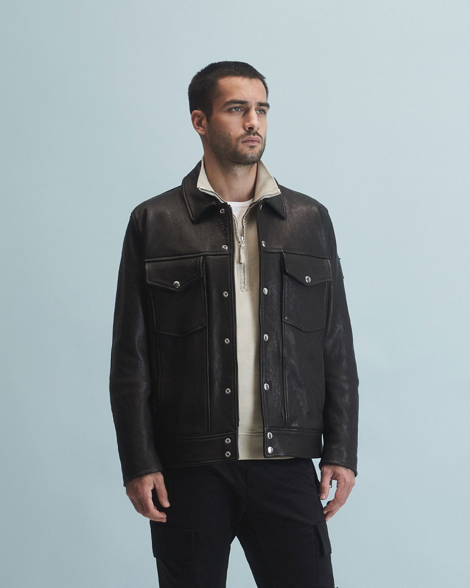 Mann mit kurzem dunklem Haar trägt schwarze Lederjacke, beigen Troyer mit Reißverschluss und schwarze Hose vor hellblauem Hintergrund.