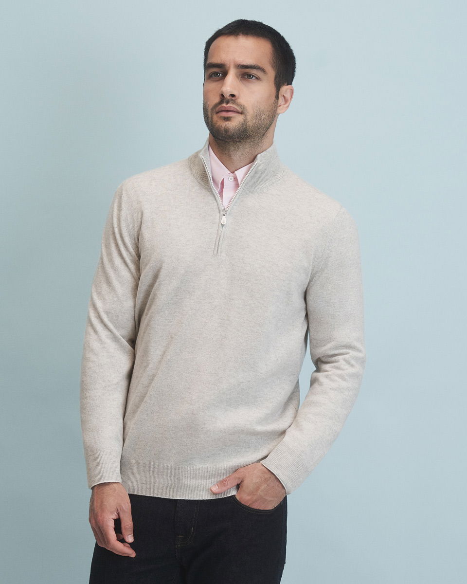 Mann mit kurzem Haar trägt hellgrauen Troyer mit Reißverschluss, rosafarbenes Hemd und schwarze Hose vor hellblauem Hintergrund.