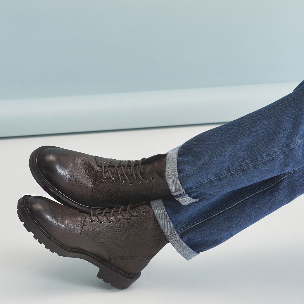 Braune Herren-Schnürboots aus Leder, getragen zu hochgekrempelten blauen Jeans vor hellem Hintergrund.