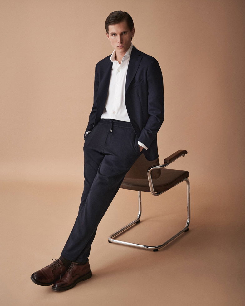 Mann lehnt am Stuhl, trägt navyblaues Sakko, weiße Hemd, Jogpants und braune Lederschuhe vor beigem Hintergrund.