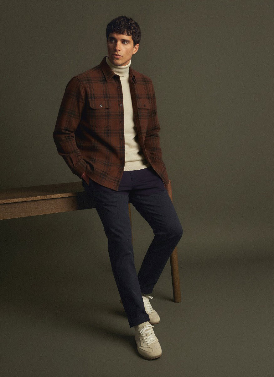 Mann lehnt am Tisch, trägt braunes kariertes Overshirt, weißen Rollkragenpullover, navy Hose und weiße Sneaker.