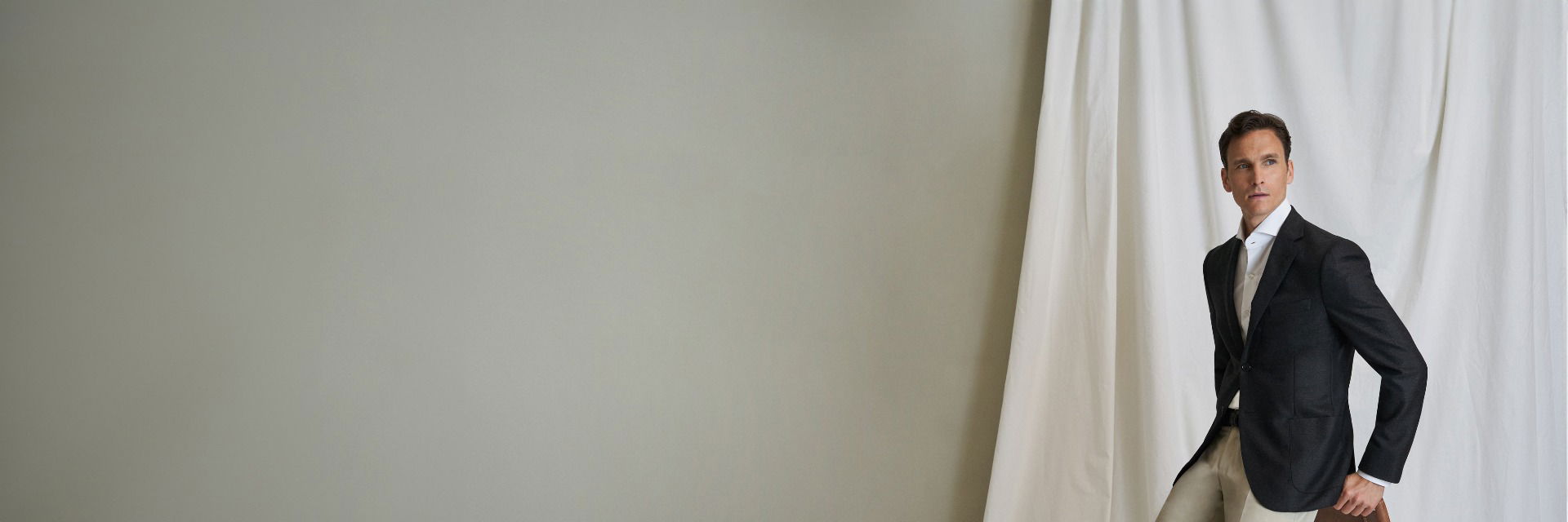 Mann lehnt an Stuhl, trägt schwarzes Sakko, weißes Hemd und beige Chinohose vor hellem Hintergrund mit weißem Vorhang. Mann lehnt an Stuhl, trägt schwarzes Sakko, weißes Hemd und beige Chinohose vor hellem Hintergrund mit weißem Vorhang.