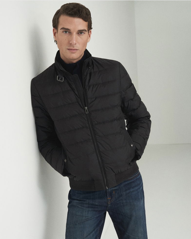 Junger Mann lehnt an weißer Wand, trägt schwarze Daunenjacke, schwarzen Rollkragenpullover und blaue Jeans.