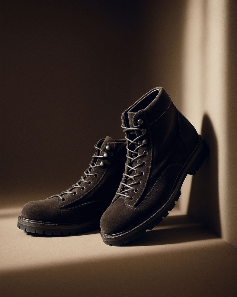 Dunkelbraune Herren-Schnürstiefel aus Veloursleder mit Gummisohle, stehend an einer beige Wand, im warmen Licht. Dunkelbraune Herren-Schnürstiefel aus Veloursleder mit Gummisohle, stehend an einer beige Wand, im warmen Licht.