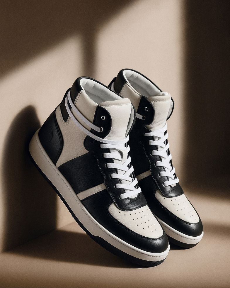 Schwarz-weiße High-Top-Sneaker für Herren, aus Leder, mit weißen Schnürsenkeln, an eine Wand gelehnt im Licht- und Schatten-Spiel. Schwarz-weiße High-Top-Sneaker für Herren, aus Leder, mit weißen Schnürsenkeln, an eine Wand gelehnt im Licht- und Schatten-Spiel.
