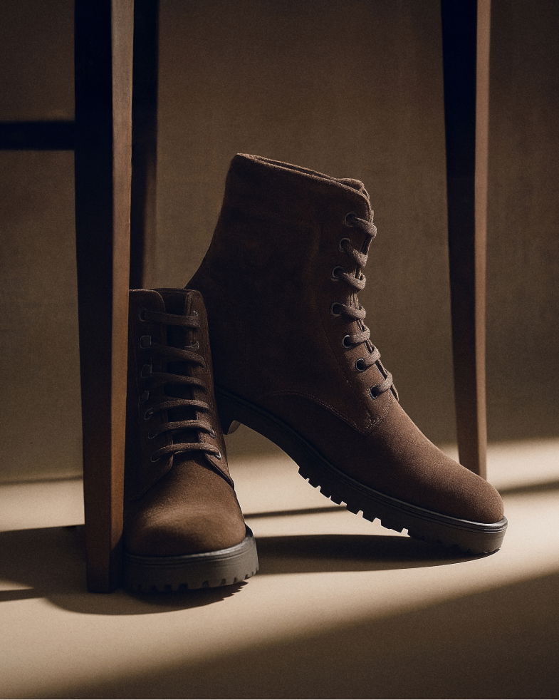 Braune Herren-Schnürboots aus Veloursleder, an ein Stuhlbein gelehnt, mit profillierter schwarzer Sohle, im warmen Licht. Braune Herren-Schnürboots aus Veloursleder, an ein Stuhlbein gelehnt, mit profillierter schwarzer Sohle, im warmen Licht.