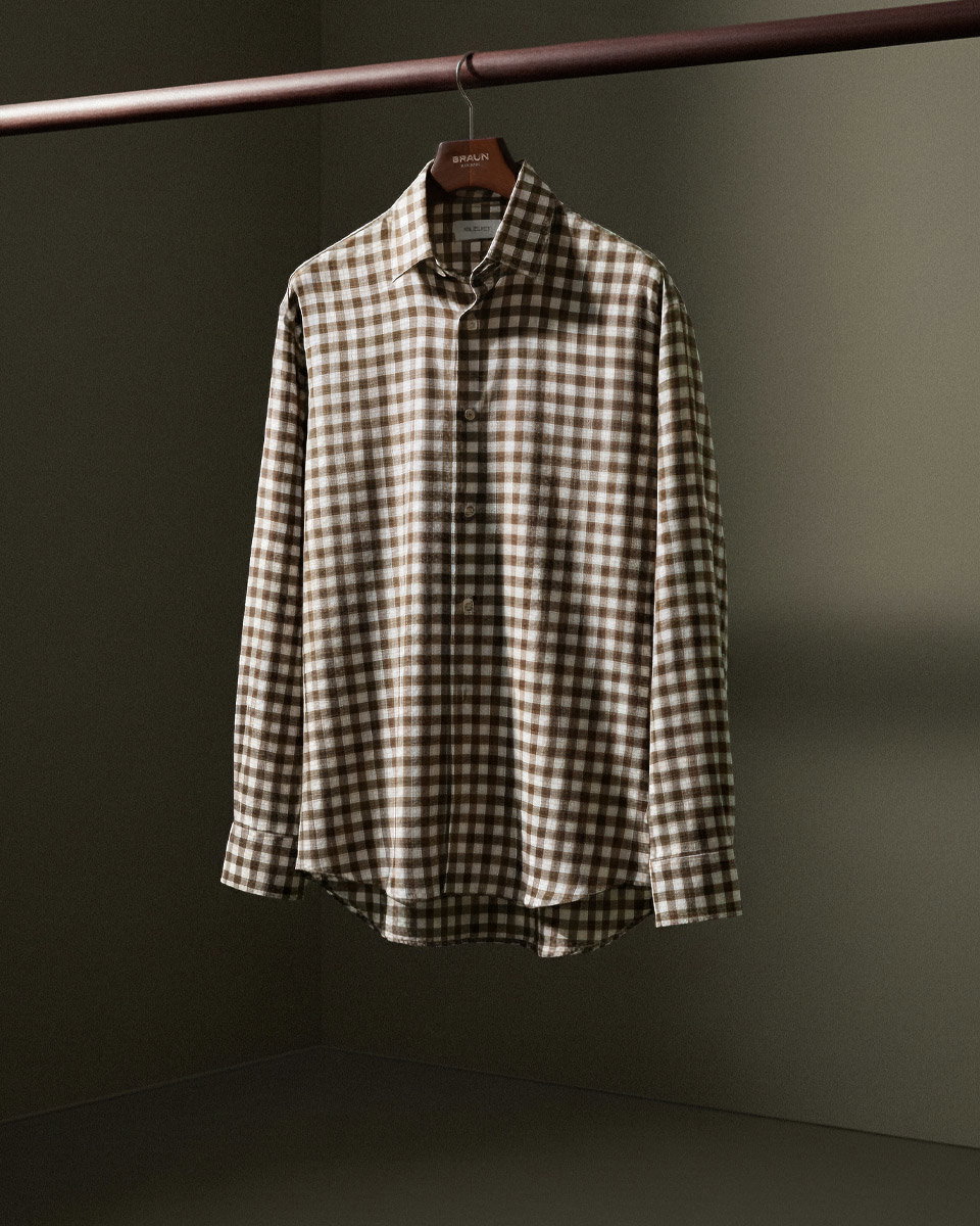 Braun-weiß kariertes Flanell-Herrenhemd, an einer Holzstange auf einem BRAUN-Kleiderbügel hängend, vor dunklem Hintergrund.