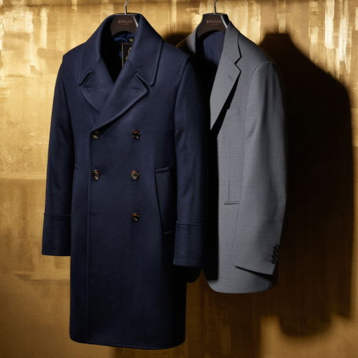 Zweireihiger navy Herren-Wollmantel und graues Sakko hängen nebeneinander an einer Kleiderstange vor goldenem Hintergrund. Zweireihiger navy Herren-Wollmantel und graues Sakko hängen nebeneinander an einer Kleiderstange vor goldenem Hintergrund.