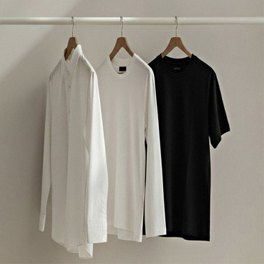 Drei Kleidungsstücke an einer weißen Stange: weißes Hemd, weißes Longsleeve und schwarzes T-Shirt, alle auf Holzbügeln.