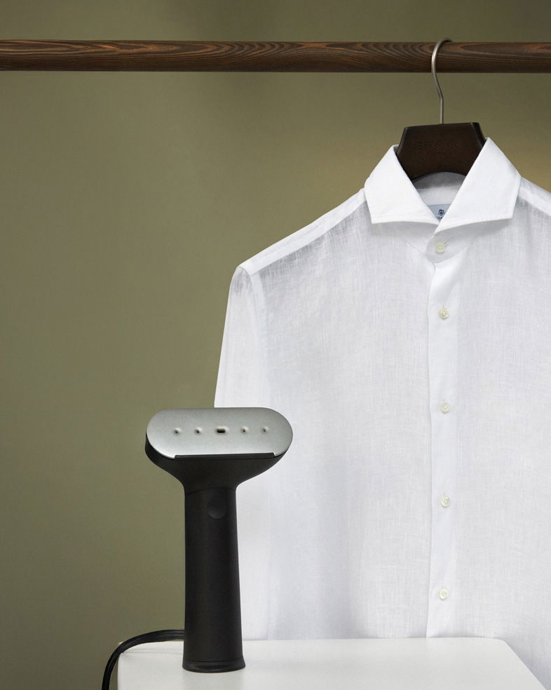 Weißes Hemd an Holzbügel hängend, davor schwarzer Handsteamer auf weißem Untergrund vor beige-grünem Hintergrund.