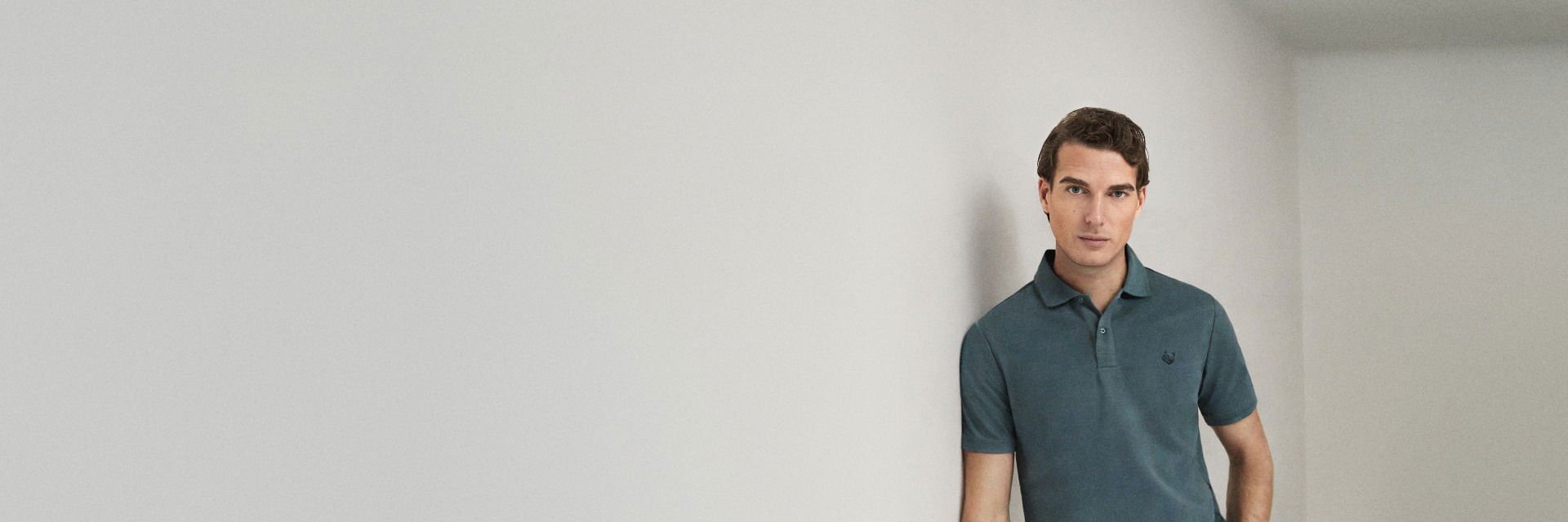 Mann lehnt an weißer Wand, trägt petrolfarbenes Poloshirt, hat kurze braune Haare und schaut direkt in die Kamera. Mann lehnt an weißer Wand, trägt petrolfarbenes Poloshirt, hat kurze braune Haare und schaut direkt in die Kamera.