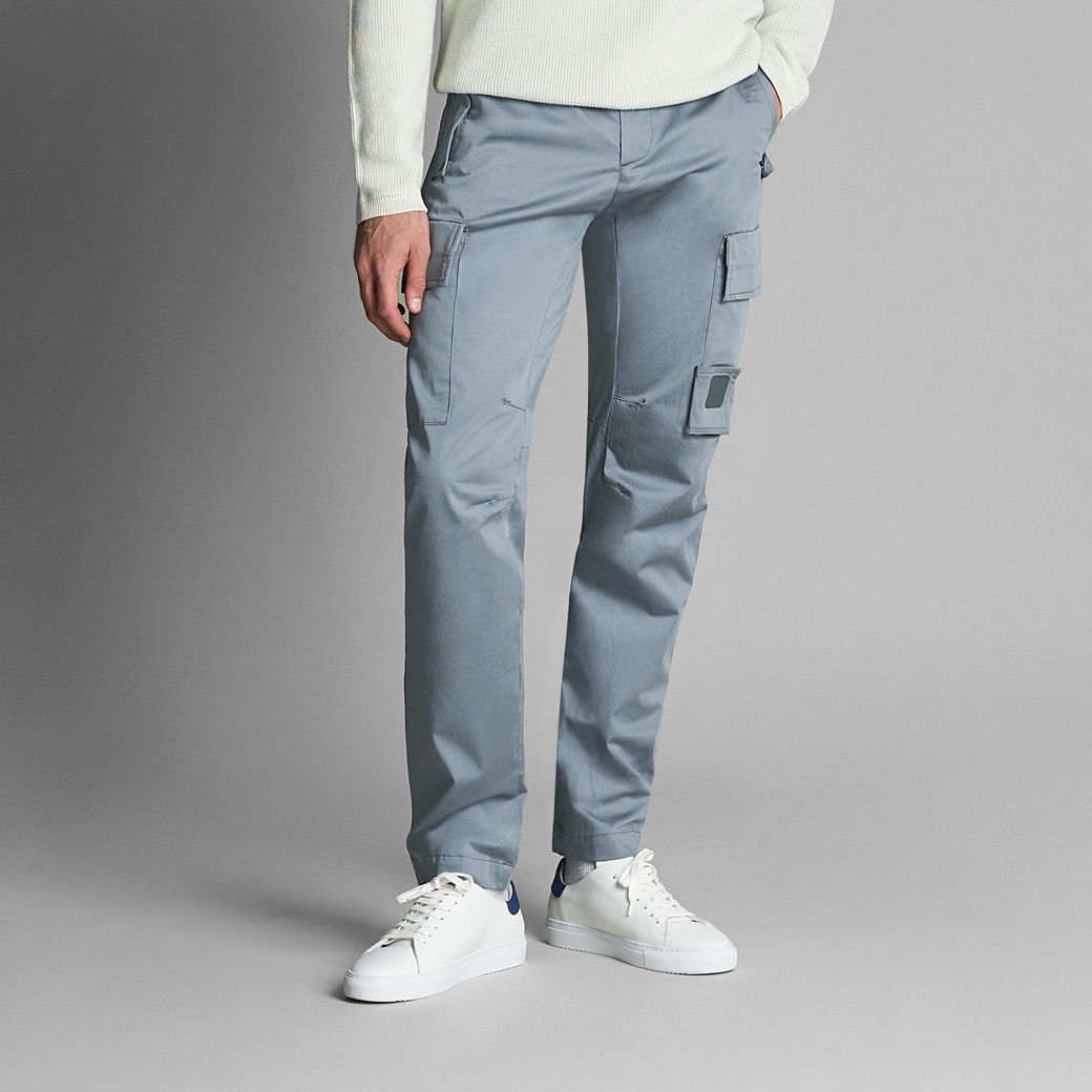 Herren tragen silbergraue Cargohose mit Taschen, weiße Sneaker und hellgrünen Pullover, vor grauem Hintergrund.