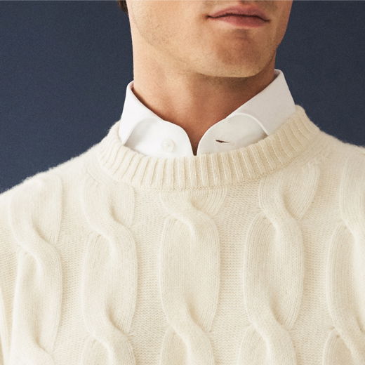 Mann trägt cremefarbenen Zopfmuster-Strickpullover über weißem Hemd, Ausschnitt von Hals bis Brust sichtbar.