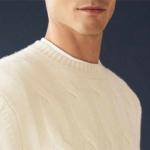 Mann trägt cremefarbenen Strick-Rundhalspullover mit Zopfmuster über weißem T-Shirt vor dunklem Hintergrund.