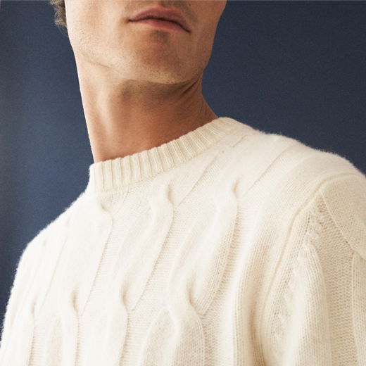 Mann trägt cremefarbenen Strick-Rundhalspullover mit Zopfmuster, Ausschnitt von Kinn bis Schulter sichtbar, dunkler Hintergrund.