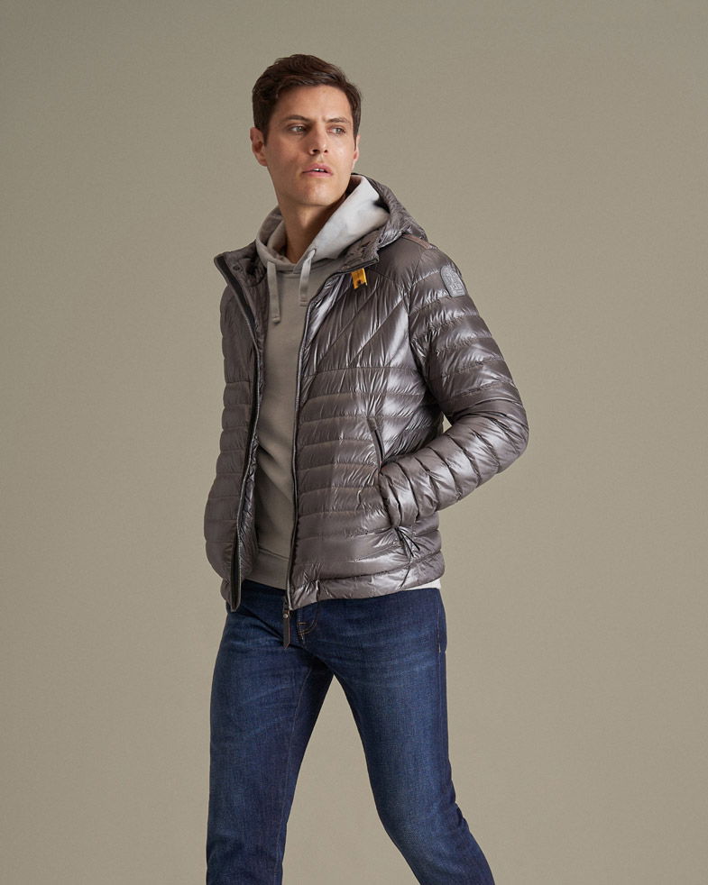 Mann trägt silbergraue Daunenjacke, hellgrauen Hoodie und dunkelblaue Jeans, steht vor beigem Hintergrund.