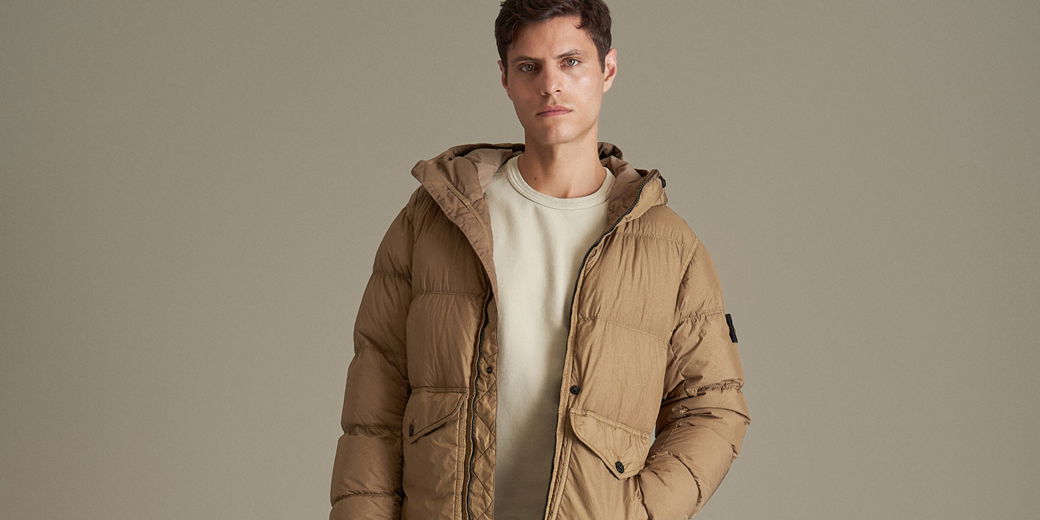 Mann mit kurzem braunem Haar trägt beige-braune Daunenjacke mit Kapuze über cremefarbenem Rundhalspullover vor beigem Hintergrund. Mann mit kurzem braunem Haar trägt beige-braune Daunenjacke mit Kapuze über cremefarbenem Rundhalspullover vor beigem Hintergrund.