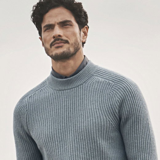 Mann mit dunklem Haar trägt hellblauen Kaschmir-Rippstrickpullover über dunkelgrauem Hemd vor hellem Hintergrund. Mann mit dunklem Haar trägt hellblauen Kaschmir-Rippstrickpullover über dunkelgrauem Hemd vor hellem Hintergrund.