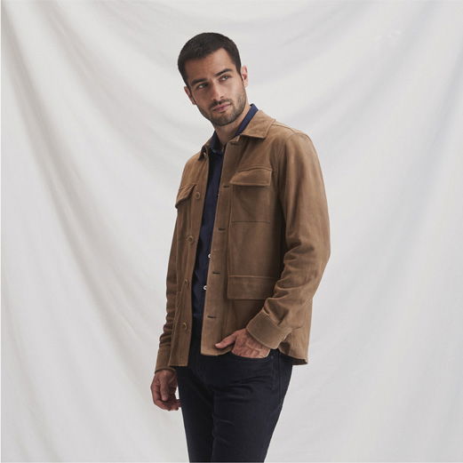 Mann trägt braunes Veloursleder-Overshirt, blaues Hemd und schwarze Hose, steht vor weißem Hintergrund, Blick nach links.