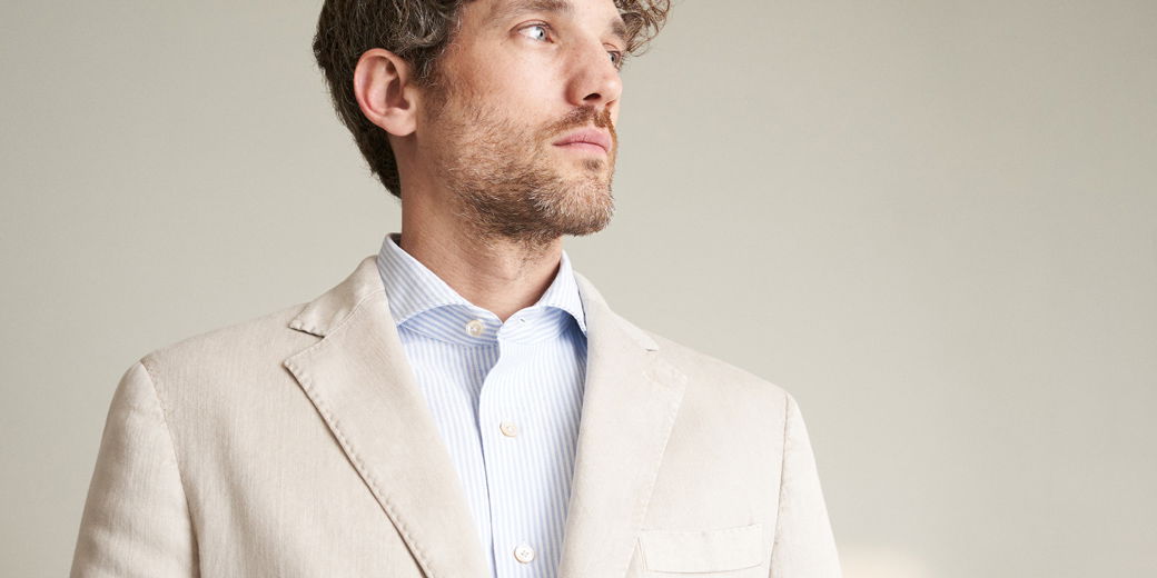 Mann mit kurzem lockigem Haar und Bart trägt beigefarbenes Sakko und weiß-blau gestreiftes Hemd, schaut nach rechts.