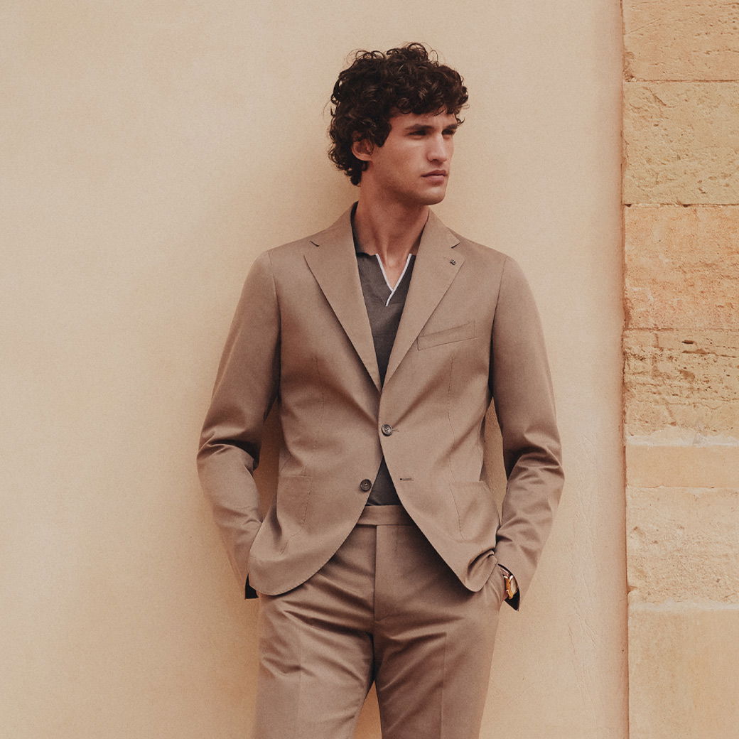 Mann mit lockigem Haar trägt beigen Anzug und braunes Poloshirt, lehnt an hellbeiger Wand mit Steinmauer rechts. Mann mit lockigem Haar trägt beigen Anzug und braunes Poloshirt, lehnt an hellbeiger Wand mit Steinmauer rechts.