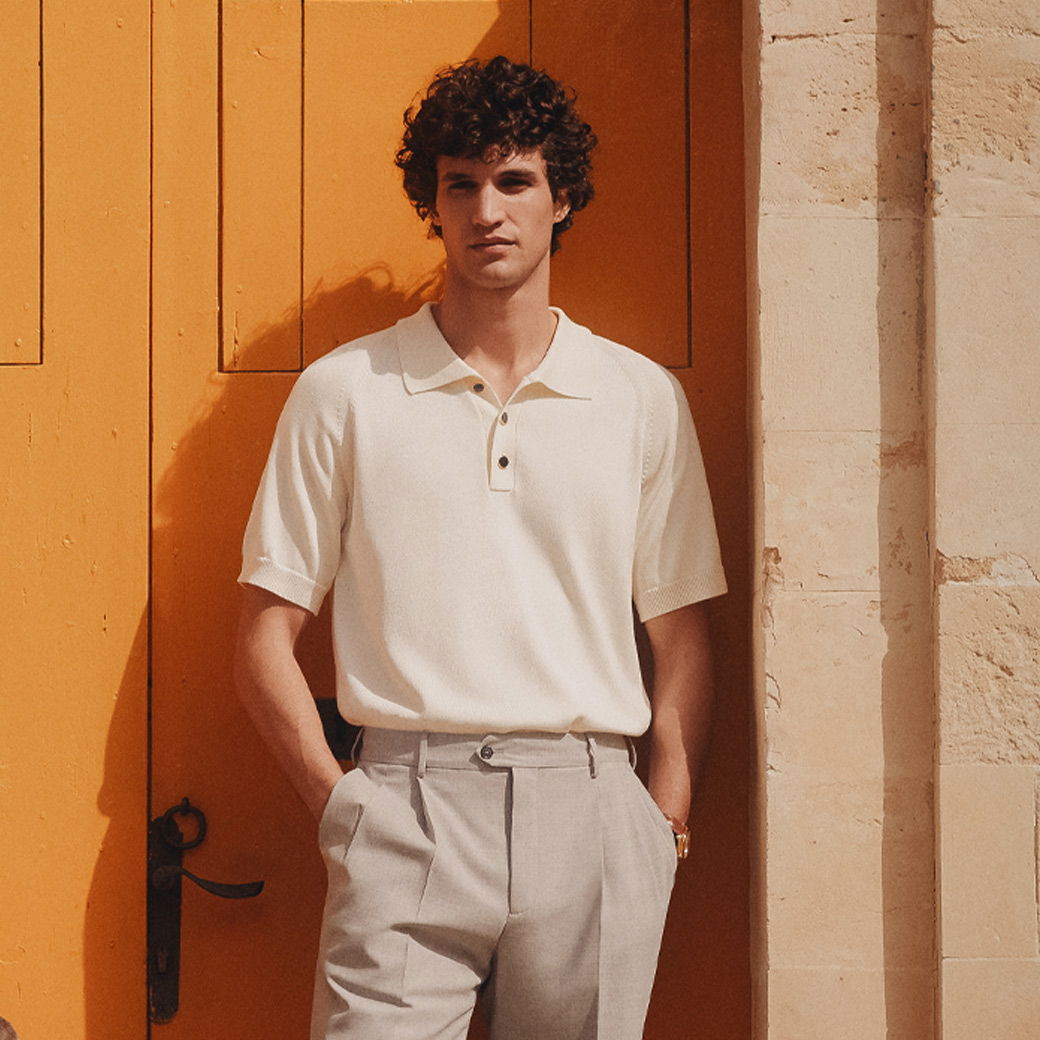 Mann mit lockigen Haaren trägt cremefarbenes Poloshirt und beige Hose, lehnt an orangefarbener Tür vor heller Wand. Mann mit lockigen Haaren trägt cremefarbenes Poloshirt und beige Hose, lehnt an orangefarbener Tür vor heller Wand.