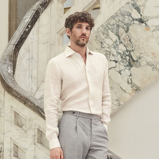 Mann mit lockigem Haar trägt cremefarbenes Leinenhemd und hellgraue Hose, steht vor marmorverkleideter Treppe.