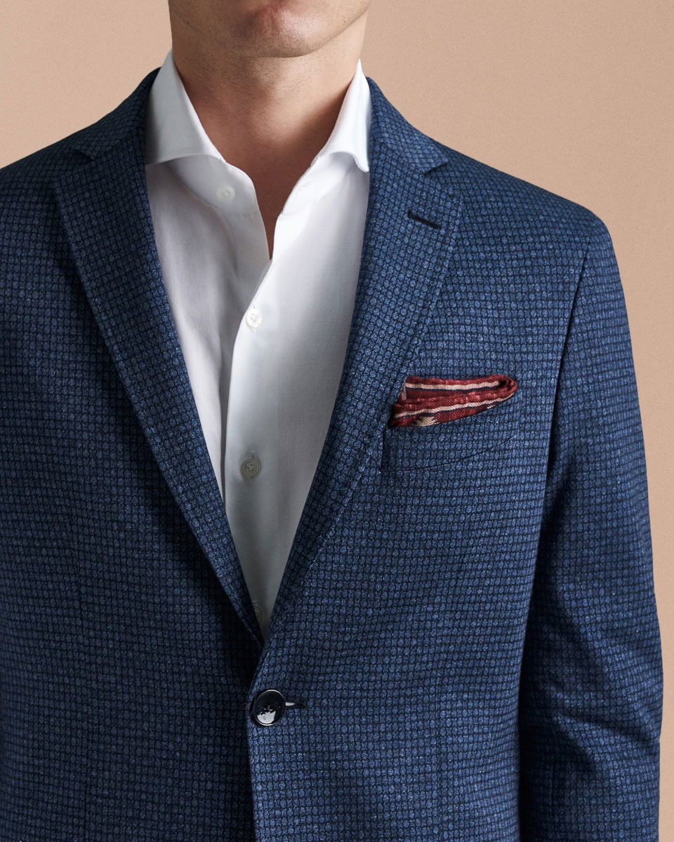 Herrenoberkörper mit dunkelblauem, gemustertem Sakko, weißem Hemd und rotem, gemustertem Einstecktuch vor beigem Hintergrund. Herrenoberkörper mit dunkelblauem, gemustertem Sakko, weißem Hemd und rotem, gemustertem Einstecktuch vor beigem Hintergrund.