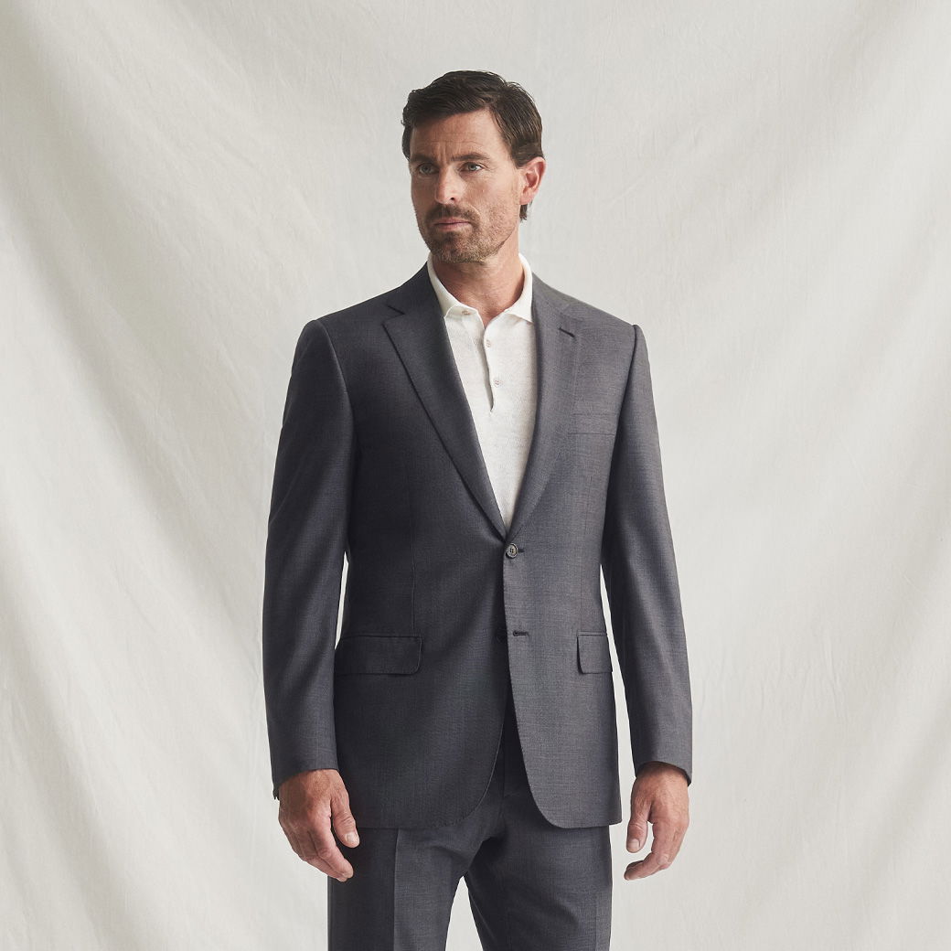 Mann trägt dunkelgrauen Anzug mit weißem Kurzarm-Strickpolo, steht vor hellem einfarbigem Hintergrund.