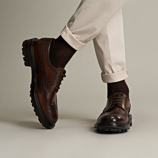 Beine mit beiger Herrenhose, hochgekrempelt, braune Vollbrogue-Lederschuhe und dunkle Socken vor neutralem Hintergrund. Beine mit beiger Herrenhose, hochgekrempelt, braune Vollbrogue-Lederschuhe und dunkle Socken vor neutralem Hintergrund.