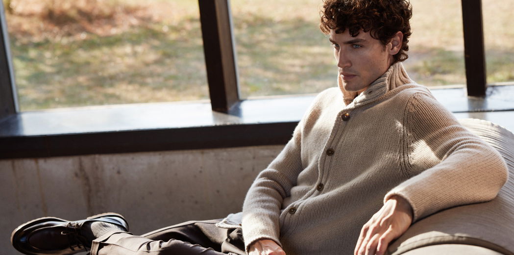 Junger Mann sitzt entspannt auf Sofa, trägt beigen Kaschmir-Cardigan mit Knöpfen, braune Hose und schwarze Schuhe. Junger Mann sitzt entspannt auf Sofa, trägt beigen Kaschmir-Cardigan mit Knöpfen, braune Hose und schwarze Schuhe.