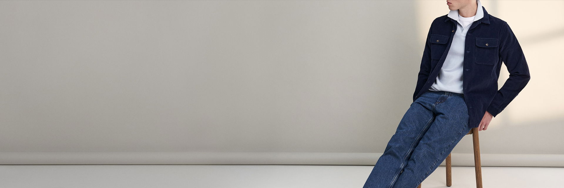 Mann sitzt auf Holzstuhl, trägt weißes Poloshirt, dunkelblaues Overshirt und blaue Jeans, vor hellem Hintergrund. Mann sitzt auf Holzstuhl, trägt weißes Poloshirt, dunkelblaues Overshirt und blaue Jeans, vor hellem Hintergrund.