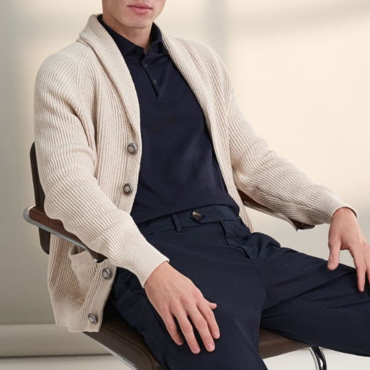 Mann sitzt auf Stuhl, trägt navy Poloshirt, beige grob gestrickten Cardigan mit Knöpfen und dunkelblaue Hose.