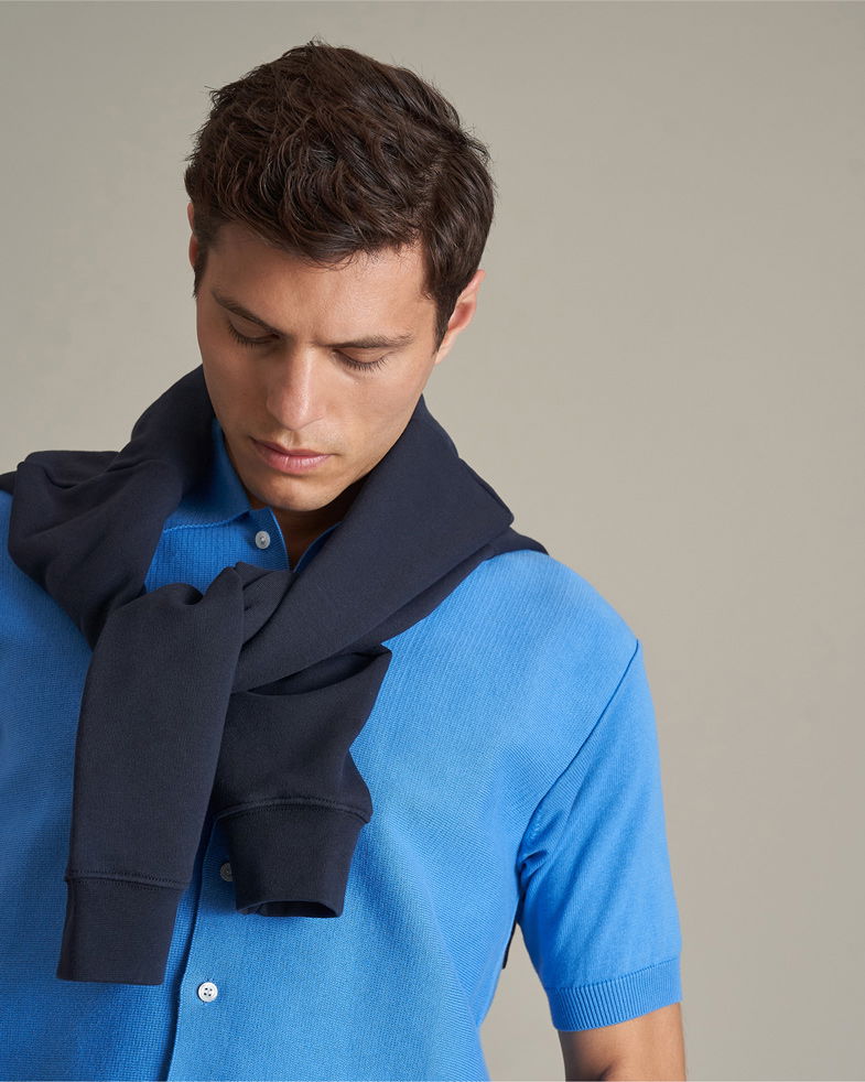 Mann mit kurzen braunen Haaren trägt blaues Strick-Kurzarmhemd, dunklen Hoodie locker um Schultern gebunden, blickt nach unten.