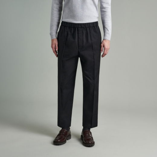 Mann trägt schwarze Loose-Fit-Hose mit elastischem Bund, grauen Pullover und braune Lederschuhe vor grauem Hintergrund. Mann trägt schwarze Loose-Fit-Hose mit elastischem Bund, grauen Pullover und braune Lederschuhe vor grauem Hintergrund.